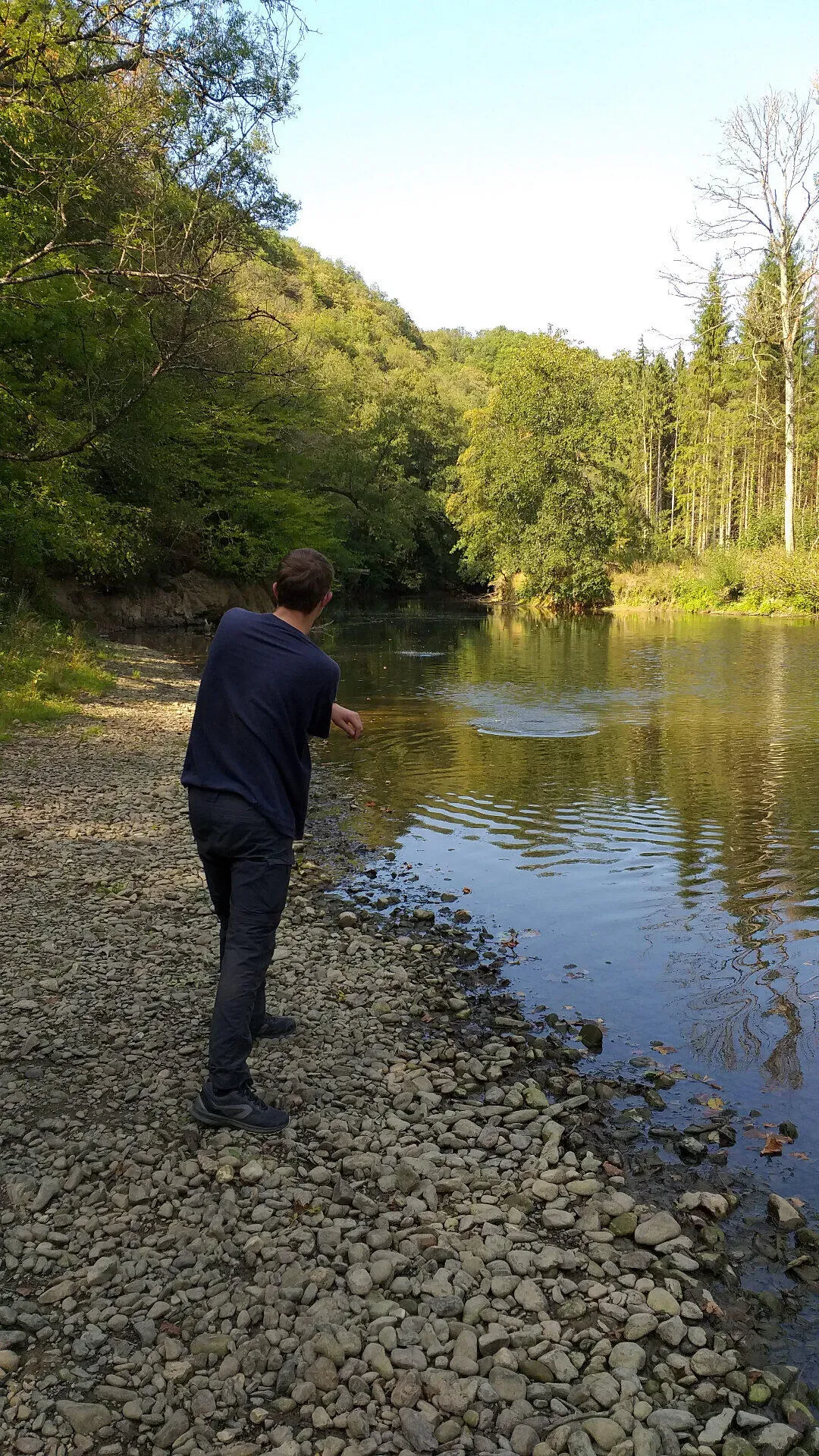 Wandelaar gooit steentjes in de Lesse vanaf kiezelstrand omringd door heuvels