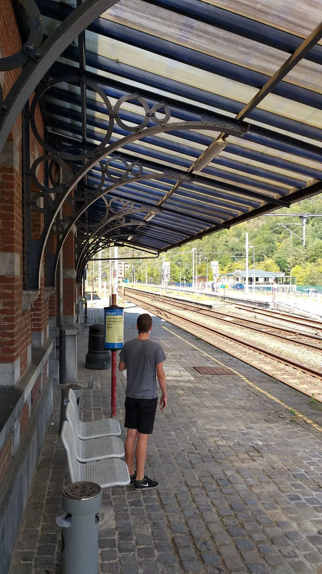 Wandelaar wacht op perron van station Houyet onder siersmeedijzeren overkapping